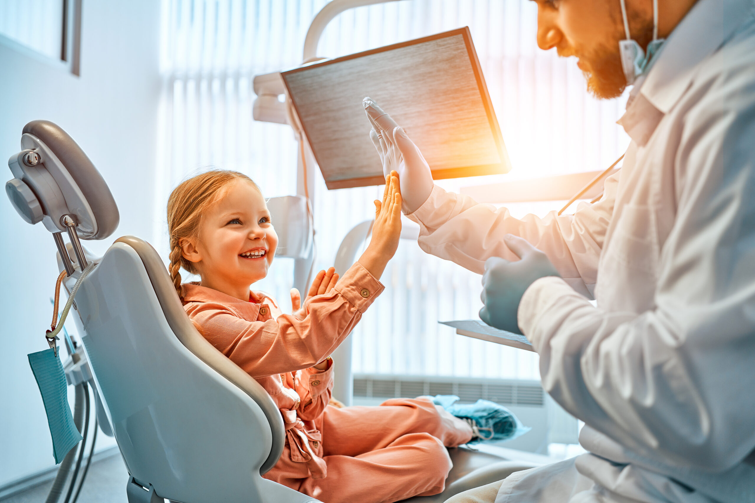 A little girl is sitting in a dentist’s chair, giving a high five to the doctor and laughing. Dental care, trust and patient care. Children’s dentistry.Sunlight.